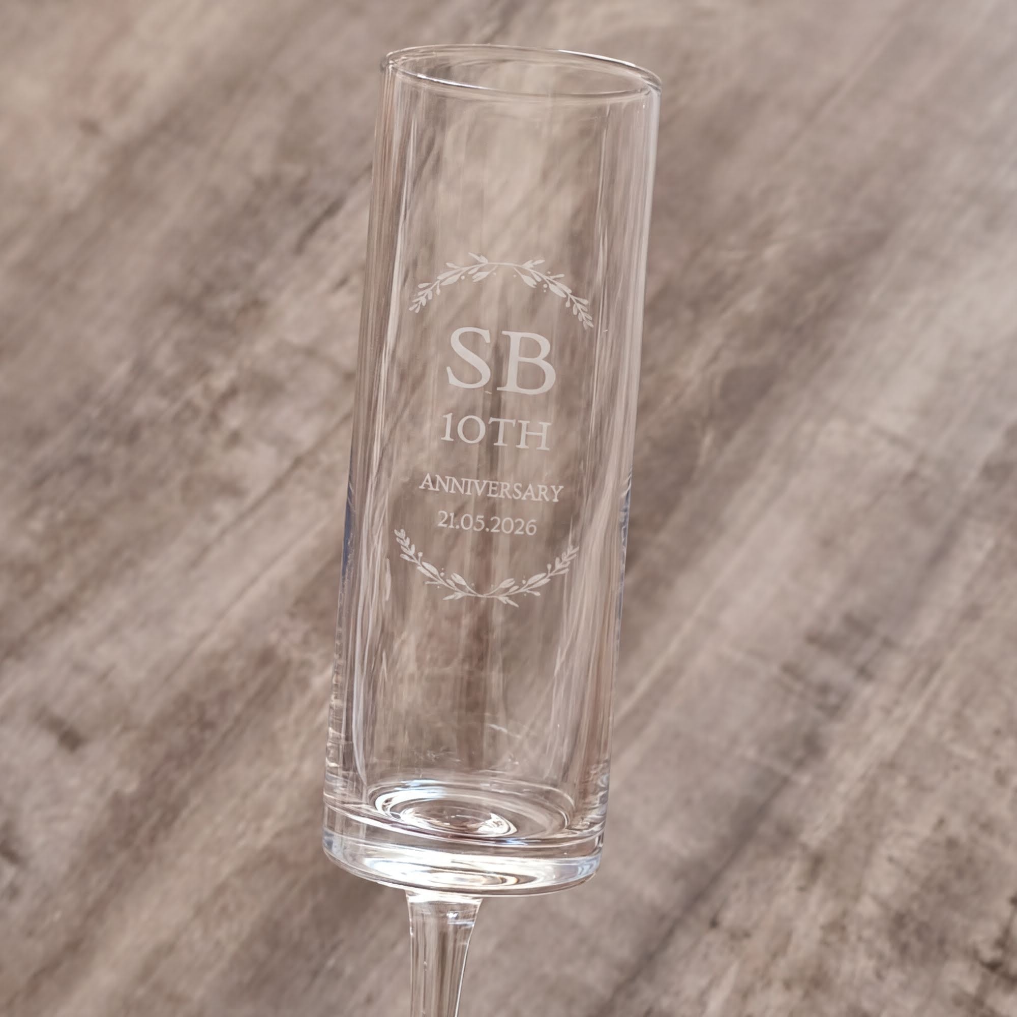A clear glass with the inscription "SB 10th ANNIVERSARY 2.05.2026" engraved on it, standing on a wooden surface.