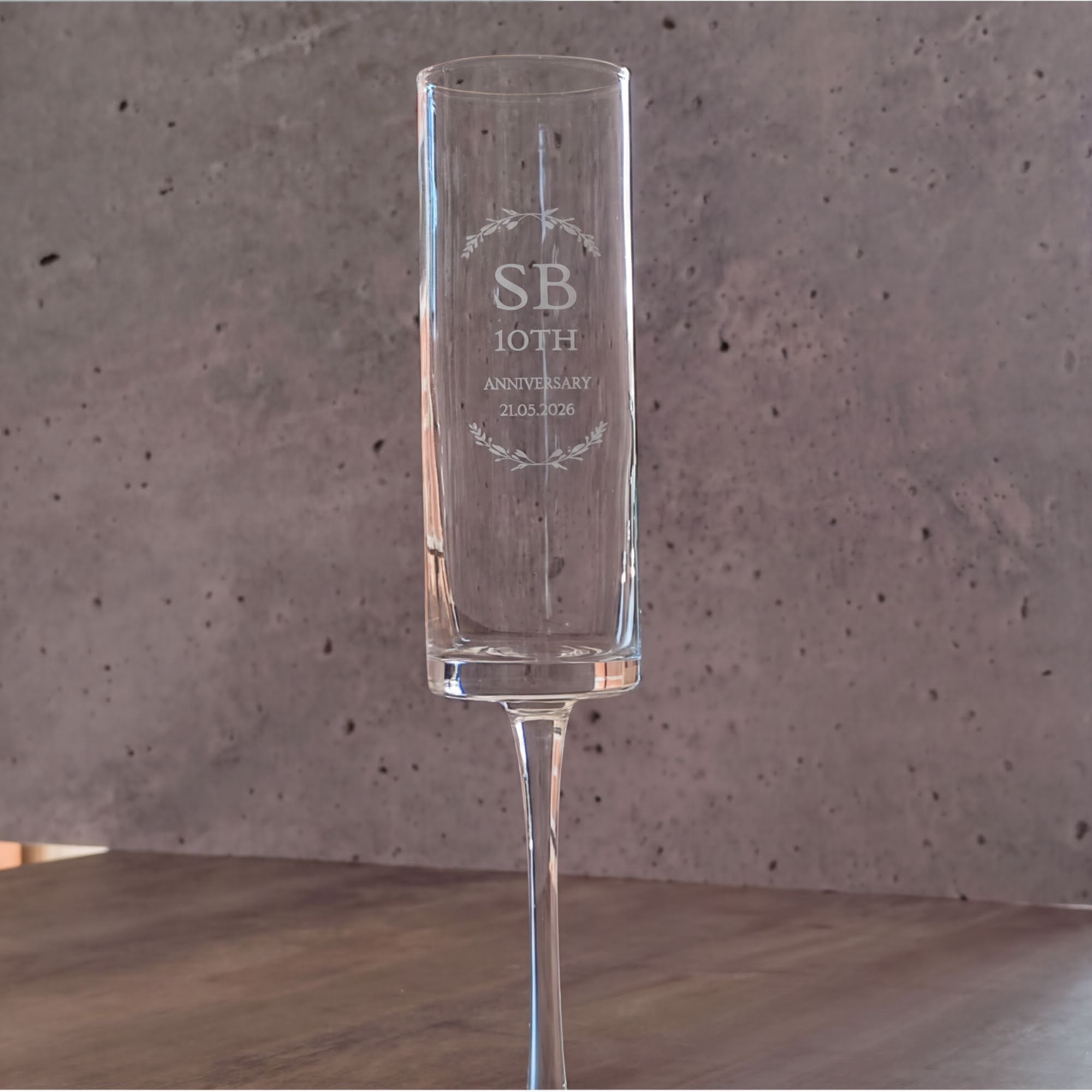 A clear glass with the inscription "SB 10th ANNIVERSARY 21.02.20" engraved on it, standing on a wooden surface against a concrete wall.