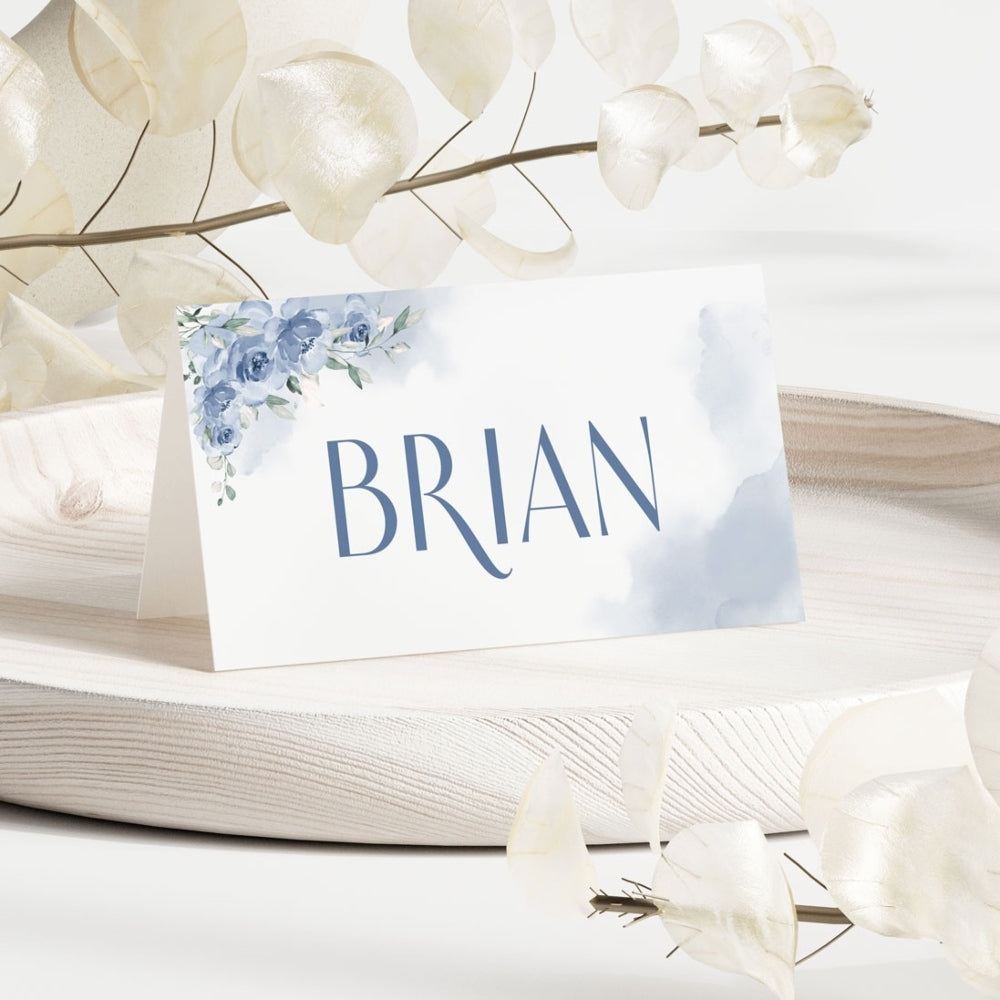 A white place card holder with a blue floral design and the name 'Brian' printed on it, displayed on a beige surface with some decorative leaves around.
