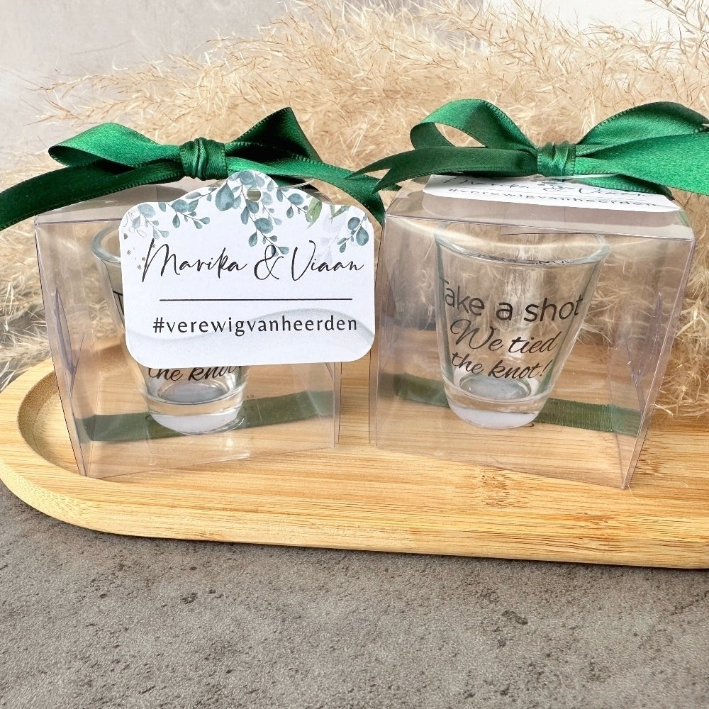 Two shot glasses in clear packaging with green ribbons on a wooden tray.