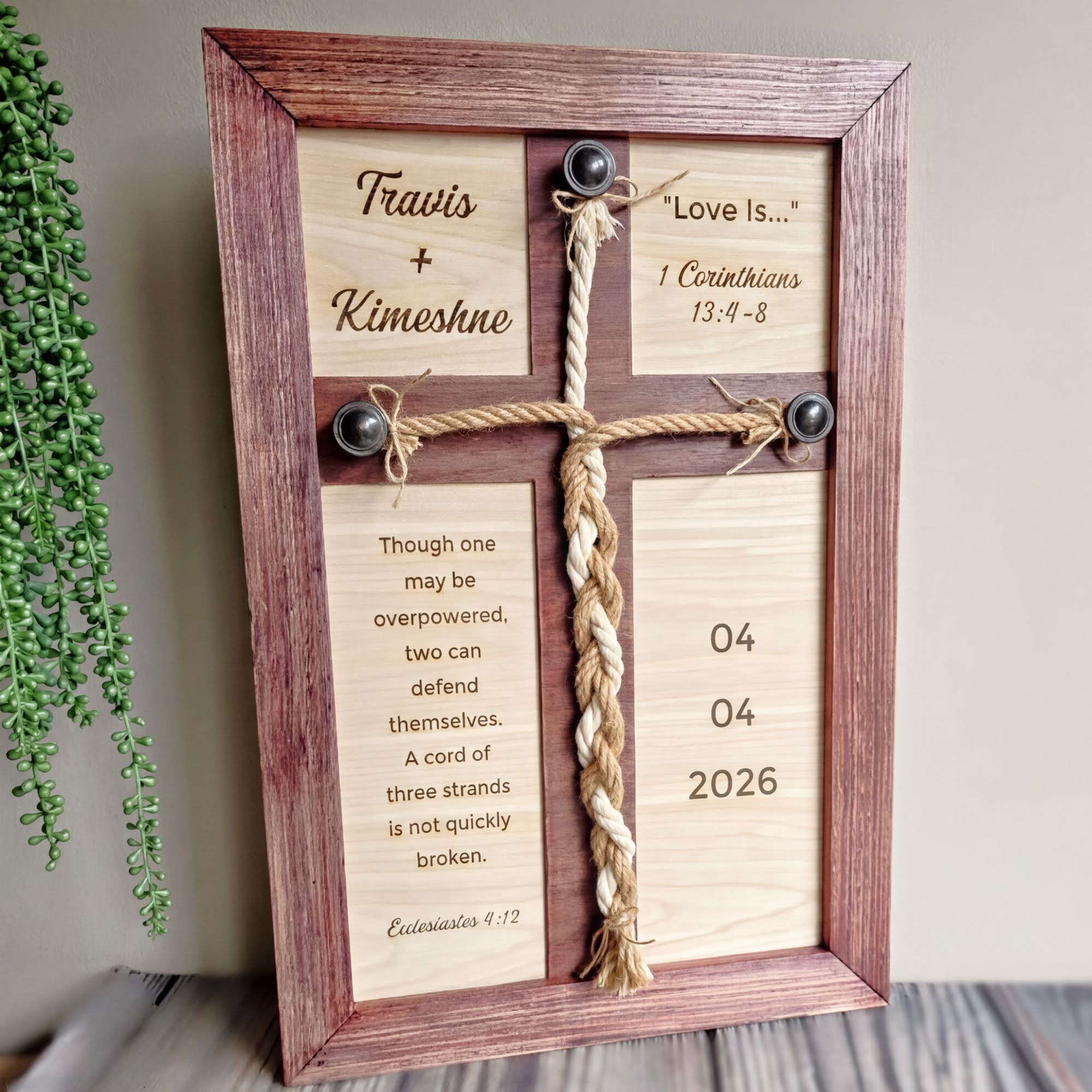 A wooden cross frame with a religious message and a date, placed on a wooden surface next to a plant.