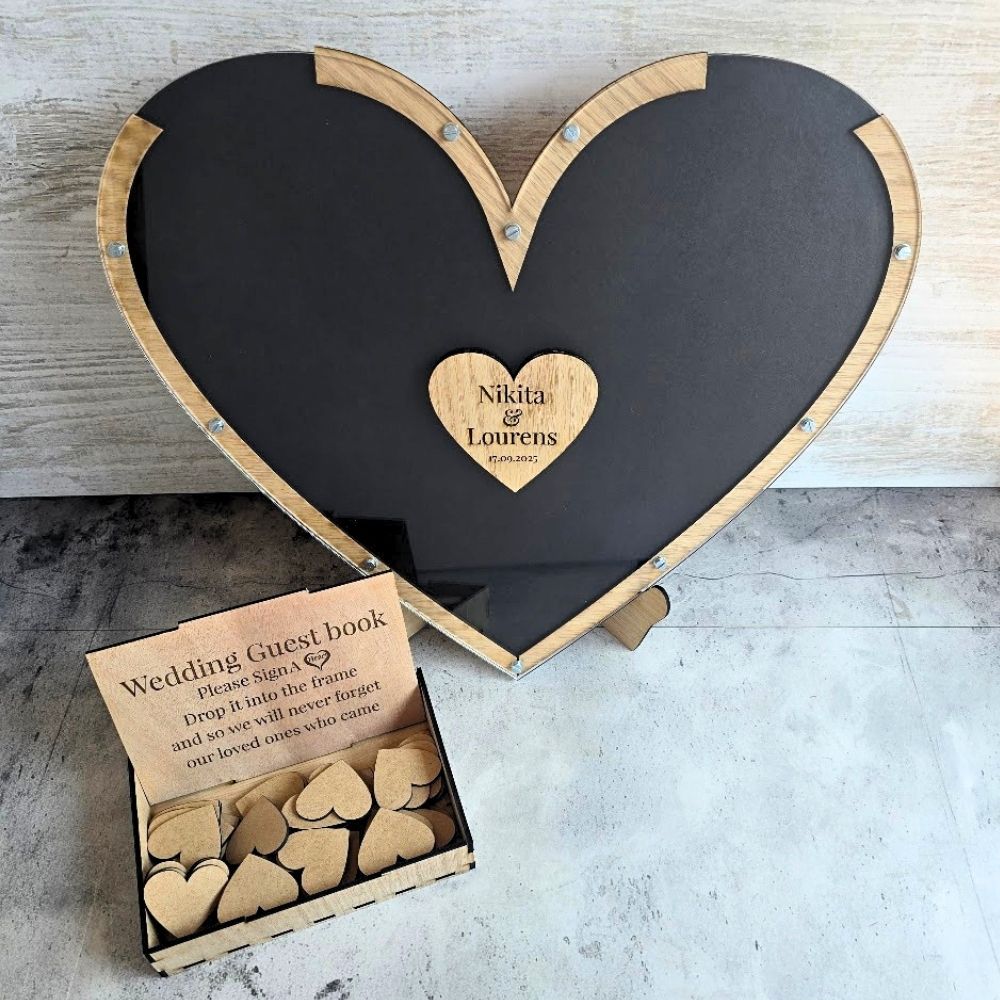 Heart-shaped wooden wedding guest book alternative with a black center panel and natural wood border, shown with an open box of small wooden heart tags on a light stone surface.