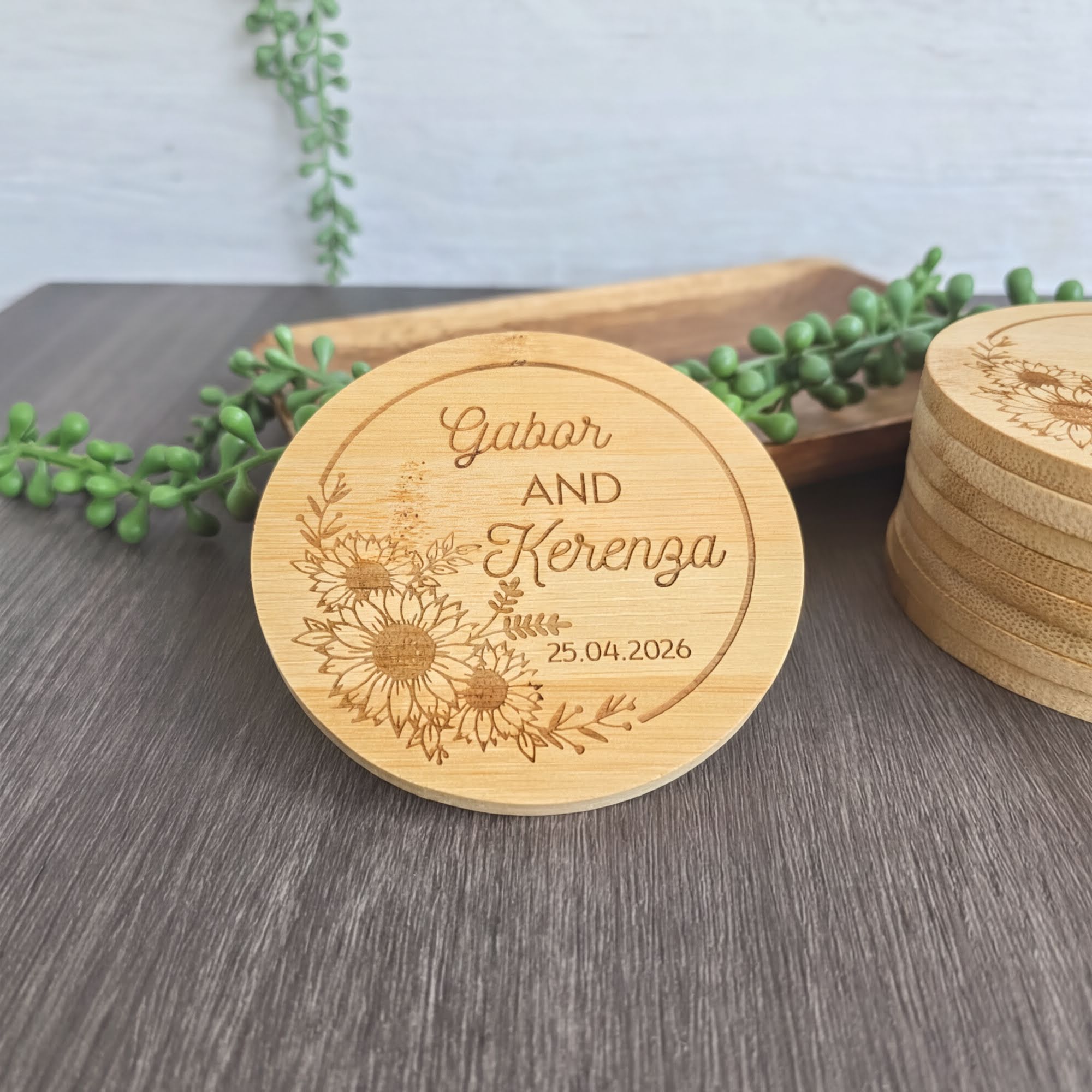 A wooden coaster with the names "Gabar and Kerenza" and the date "25.04.2026" engraved on it, along with a floral design. The coaster is placed on a wooden surface, with some green plants in the background.