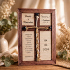 A wooden cross-shaped frame with a Bible verse and a date displayed on it, placed on a wooden surface with some greenery in the background.