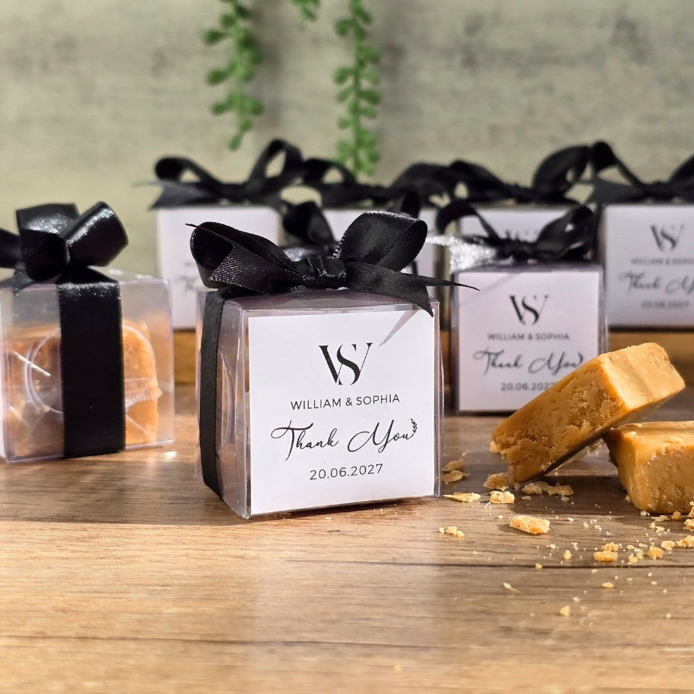 Personalized clear favour boxes wrapped in black ribbon bows, displayed on a wooden table with pieces of caramel fudge beside them. The front box is labeled with a custom monogram 'W & S' and the text 'William & Sophia Thank You.