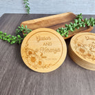 two wooden coasters with engraved text, placed on a wooden surface. The coasters are surrounded by some greenery, creating a natural and decorative setting.