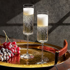 Two champagne flutes filled with champagne sit on a gold tray, accompanied by a bunch of red grapes.