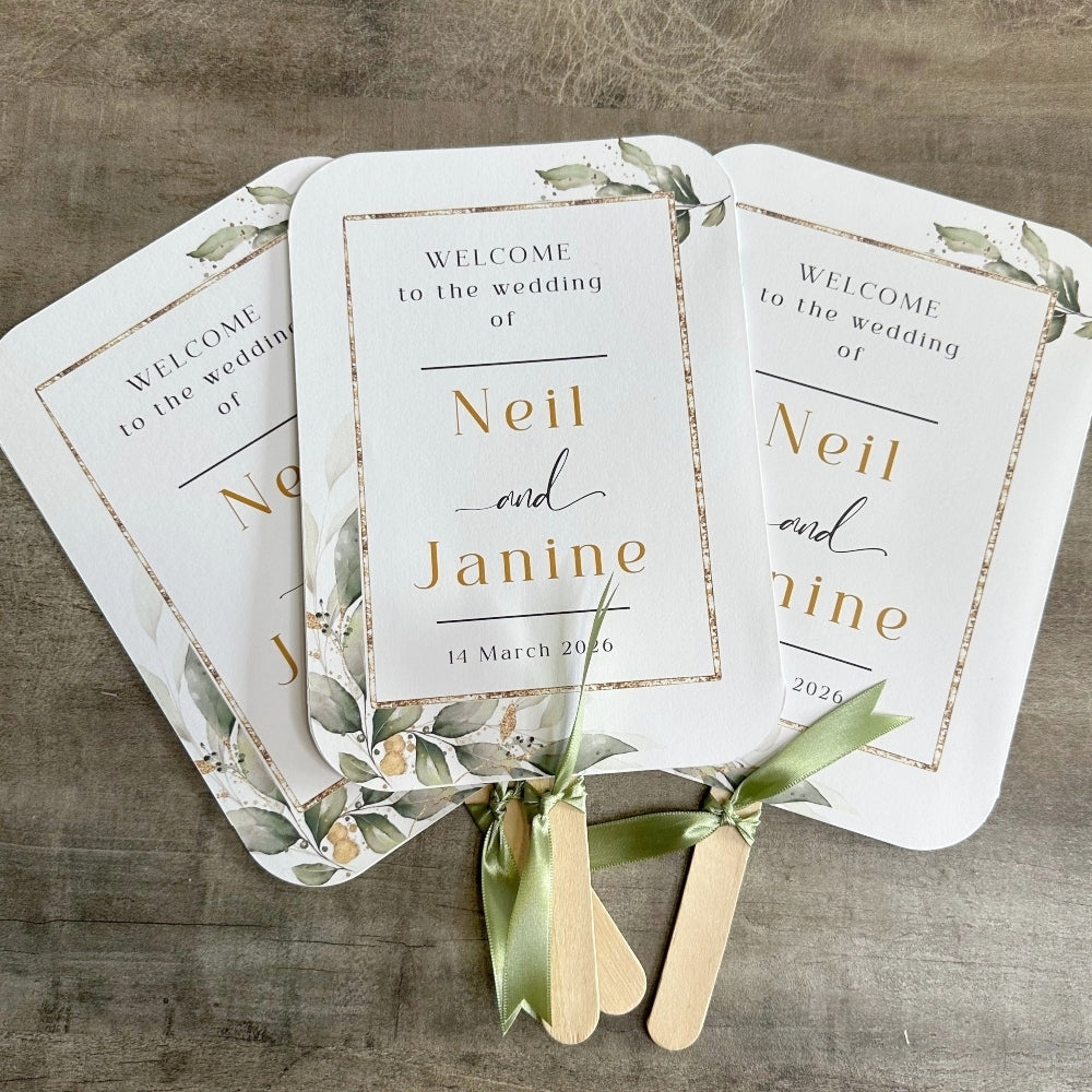 three white wedding invitations with gold leaf designs, each featuring the names "Neil and Janine" and the date "14 March 2020". The invitations are accompanied by wooden popsicle sticks with green ribbons, creating a festive and welcoming atmosphere for the wedding ceremony.