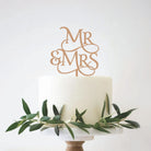 A personalized wedding cake topper featuring a laser-cut design with the initials 'MR&MRS' on top of a white cake, adorned with green leaves and a 'Mr&Mrs' sign.