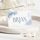 A white place card holder with a blue floral design and the name 'Brian' printed on it, displayed on a beige surface with some decorative leaves around.