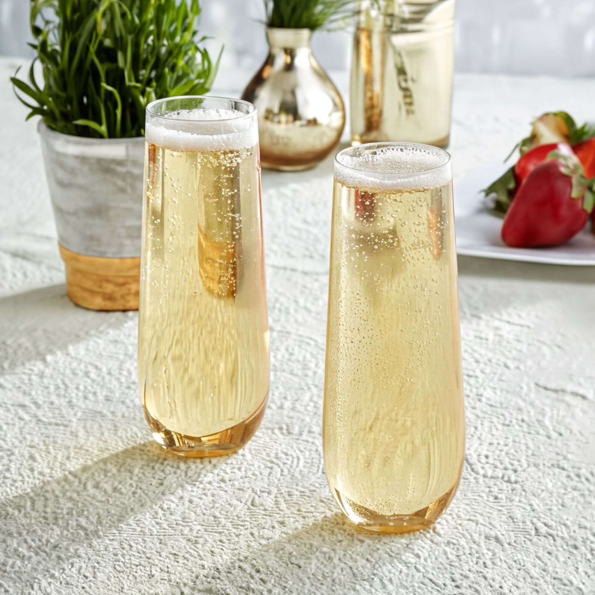 Two glasses filled with a yellowish liquid, possibly champagne or a similar beverage, placed on a table with a white tablecloth. In the background, there are some potted plants and a plate of strawberries.