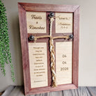 A wooden cross frame with a religious message and a date, placed on a wooden surface next to a plant.