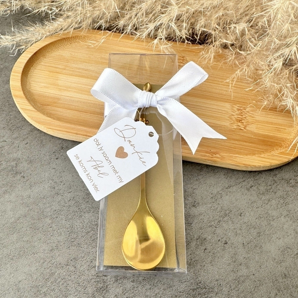 Gold spoon in a clear package with a white ribbon and tag on a stone surface.