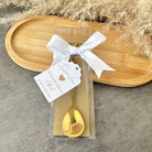 Gold spoon in a clear package with a white ribbon and tag on a stone surface.