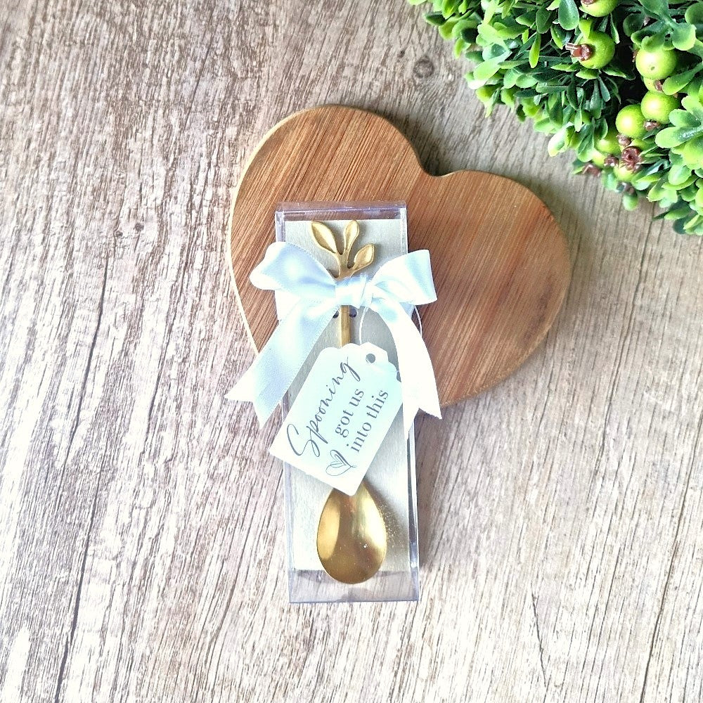 Gold spoon in a clear package with a white ribbon on a wooden heart-shaped board.