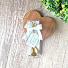 Gold spoon in a clear package with a white ribbon on a wooden heart-shaped board.