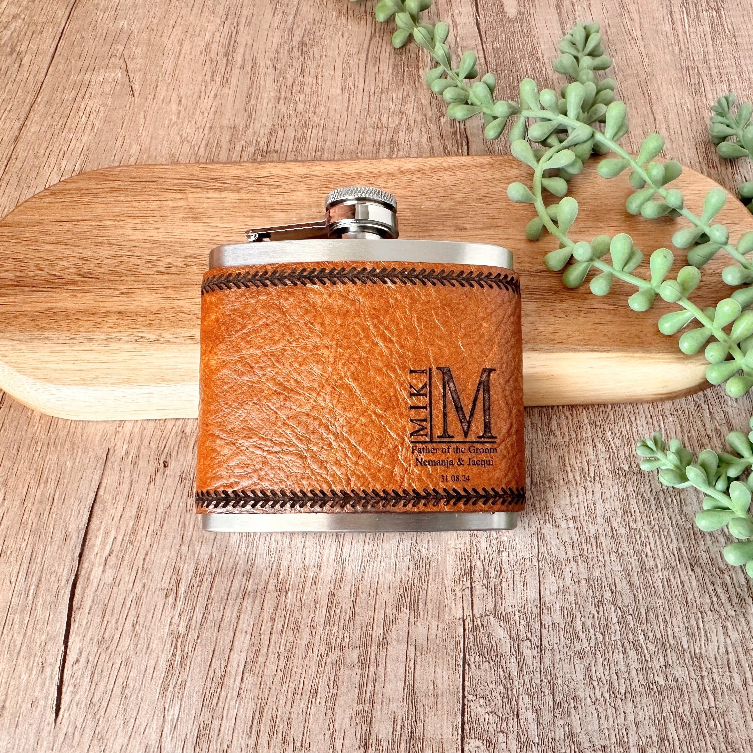 Brown leather flask with silver cap on a wooden surface with greenery