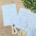 A fan program with a white background featuring text and a timeline diagram for a wedding ceremony. It is displayed with wooden sticks and decorative greenery in the background.