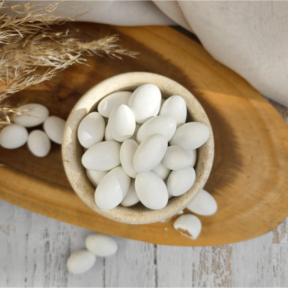 White oval sugar coated almond candies arranged in a small beige bowl on a wooden tray, with a few pieces scattered around on a light rustic surface.