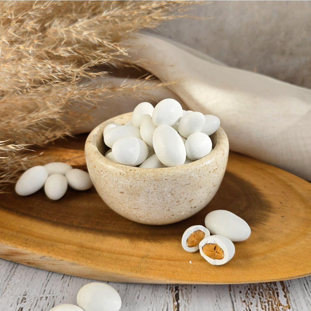 White oval-shaped sugar coasted almonds displayed in a small speckled stone bowl on a wooden tray, with some pieces broken open to show a roasted tan center, styled with dried grass and a neutral fabric background.