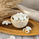 White oval-shaped sugar coasted almonds displayed in a small speckled stone bowl on a wooden tray, with some pieces broken open to show a roasted tan center, styled with dried grass and a neutral fabric background.